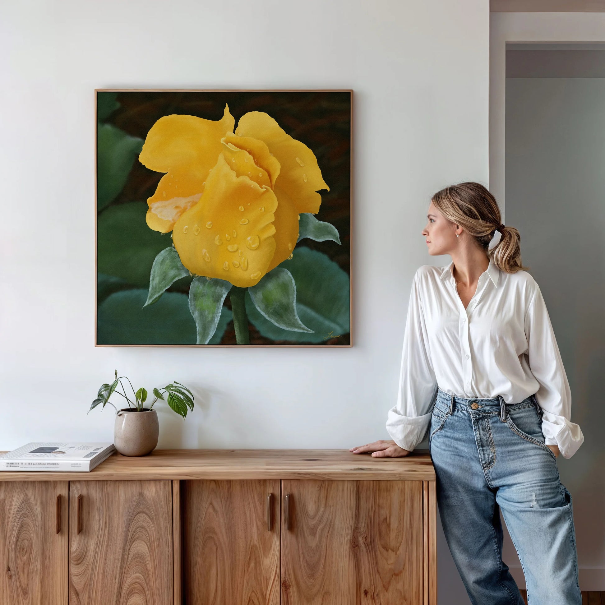 A woman in a white blouse and jeans stands by a wooden cabinet, admiring yellow rose art on a gallery-wrapped canvas with floating wood frames. A small potted plant and books rest on the cabinet beneath the painting.