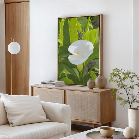 A modern living room with a light wood sideboard, beige cushions on a white sofa, potted plants, and a hyper-realistic Calla Lily painting, set against white walls and wood accents for an atmosphere of serenity and natural beauty.