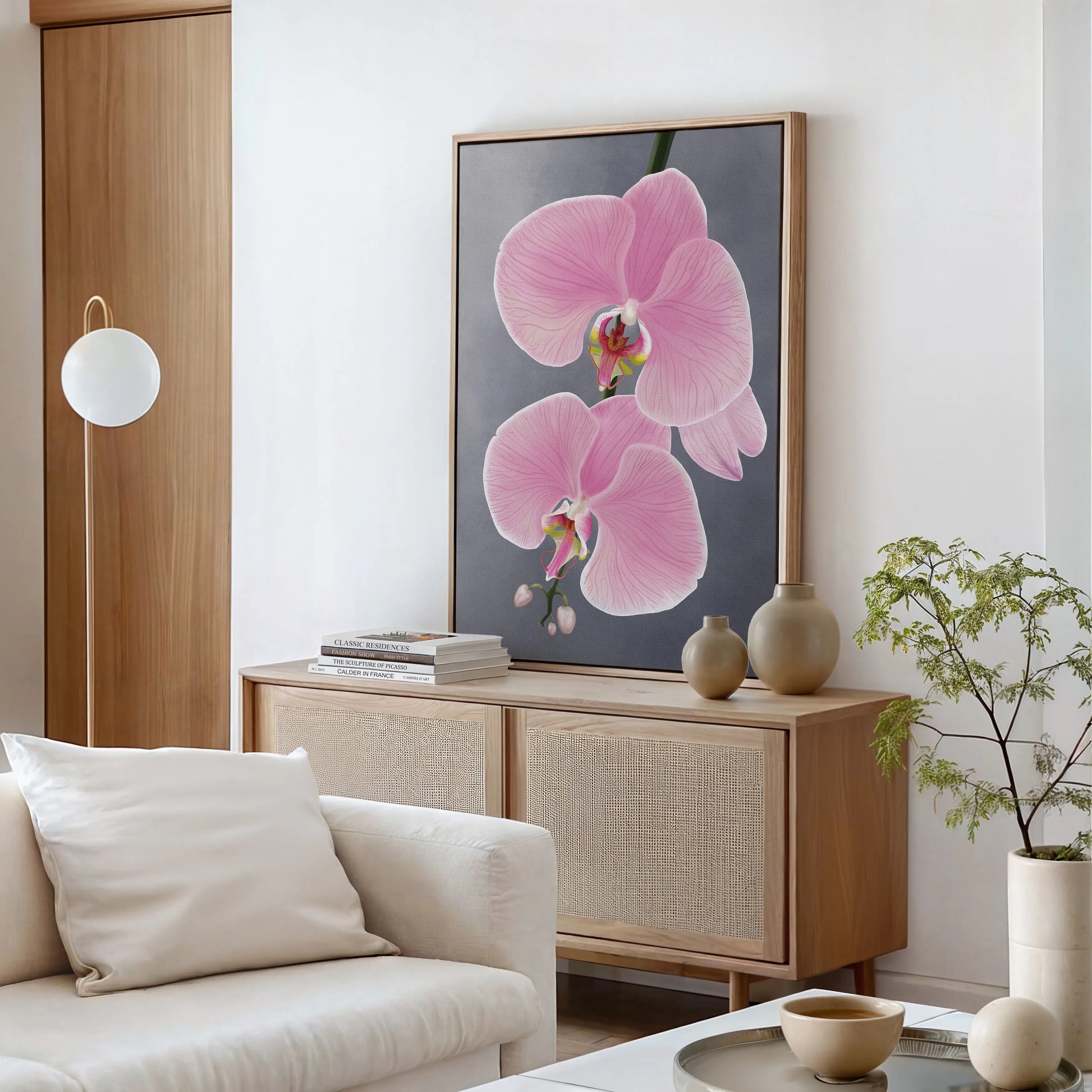 A bright modern living room with a light wood sideboard, white and beige decor, and a large gallery-wrapped canvas featuring two pink orchid flowers on a gray background. A small green plant sits on the sideboard, with a round lamp to the left.