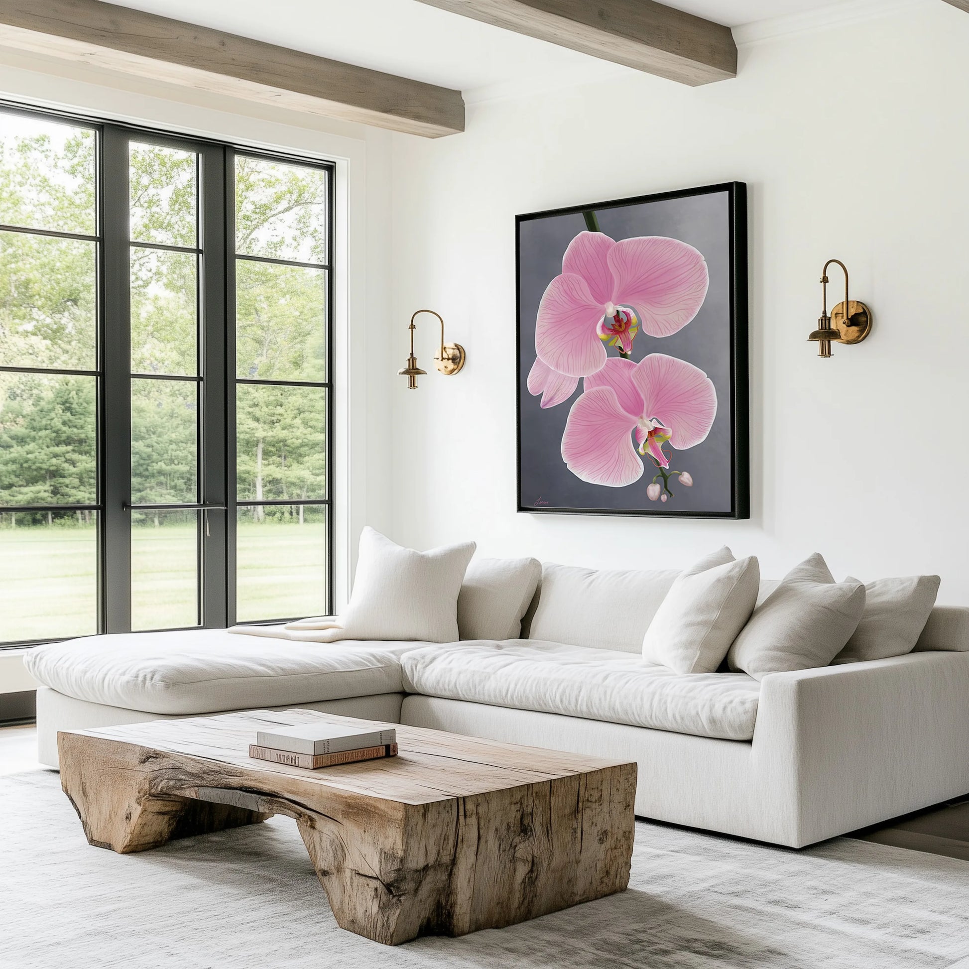 A bright, modern living room with a white sectional sofa, rustic wooden coffee table, and large windows overlooking greenery. A gallery-wrapped canvas featuring a pink orchid flower and two brass wall sconces decorate the wall above the sofa.