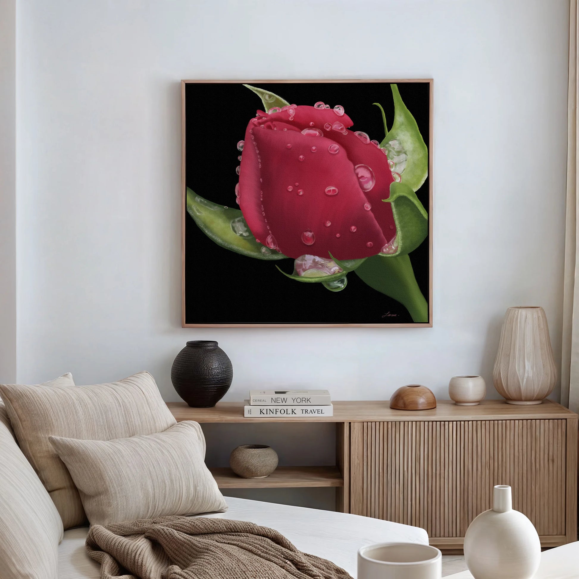 A modern living room with light neutral tones features Deeper Love, a gallery-wrapped canvas depicting a realistic red rosebud with water droplets on black, framed in wood and hung above a wooden console adorned with vases, bowls, and stacked books.