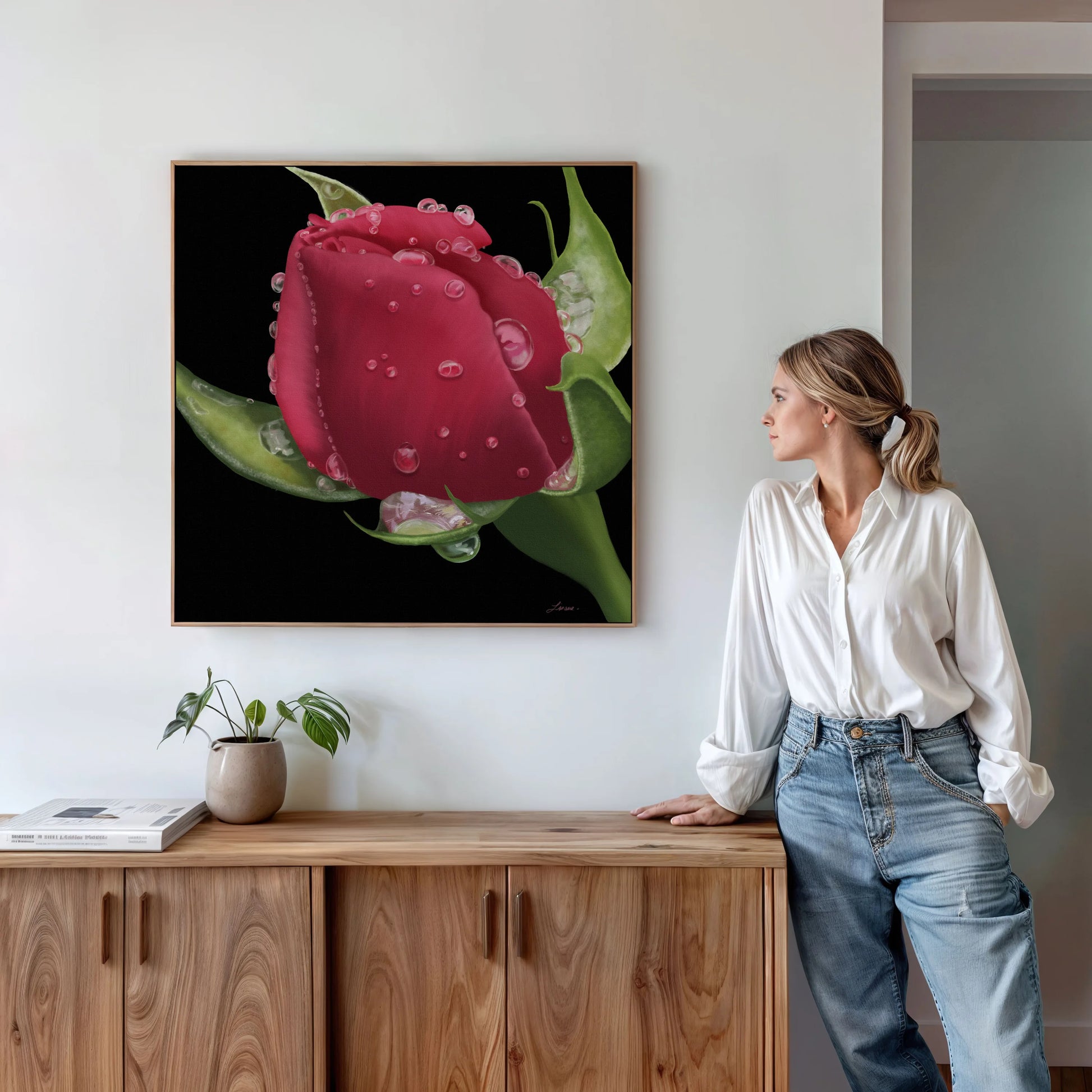 A woman in a white blouse and jeans stands by a wooden cabinet, admiring a large premium canvas painting of a red flower with vibrant color, water droplets, and green leaves. A potted plant and books accent the minimalist, modern room.