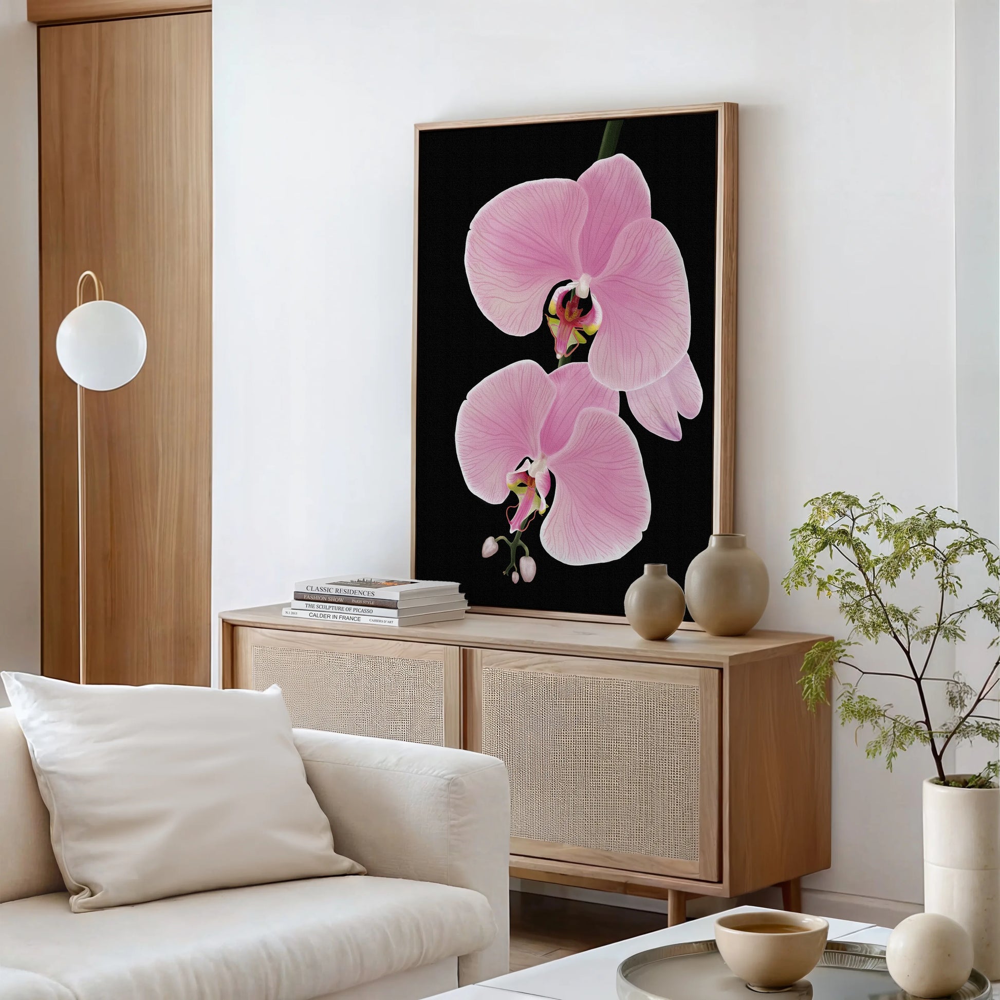 A minimalist living room with neutral tones features a light wood sideboard, white sofa, potted plant, and large gallery-wrapped canvas of orchid flowers—perfect for statement walls. A round white lamp and stacked books complete the serene look.