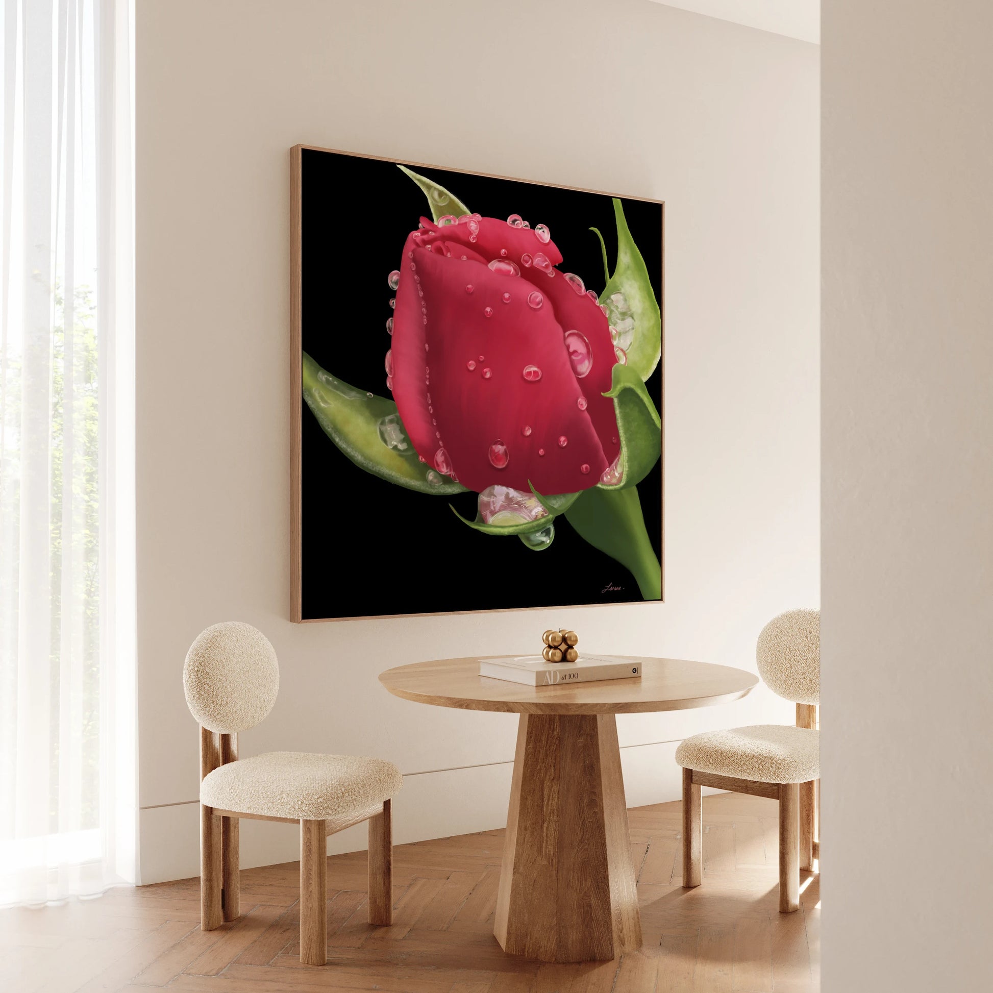 A modern, minimal dining space with two cream upholstered chairs and a round wooden table. On the wall hangs a hyperrealistic canvas of a red rosebud with water droplets in a floating wood frame, bathed in natural light from the left.