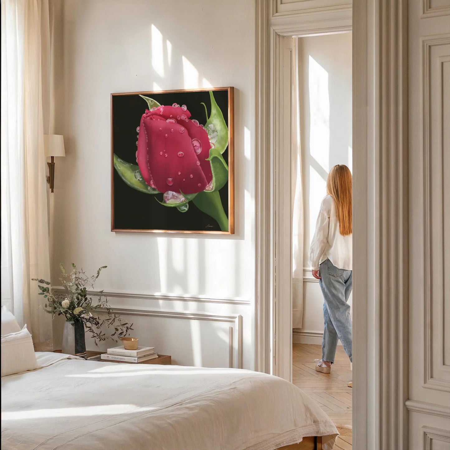 A softly lit bedroom with white walls features a bed with neutral bedding, a wooden nightstand holding a vase of white flowers, and a large hyperrealistic red rose on gallery-wrapped canvas. A person with long hair stands by a bright window in the adjoining room.