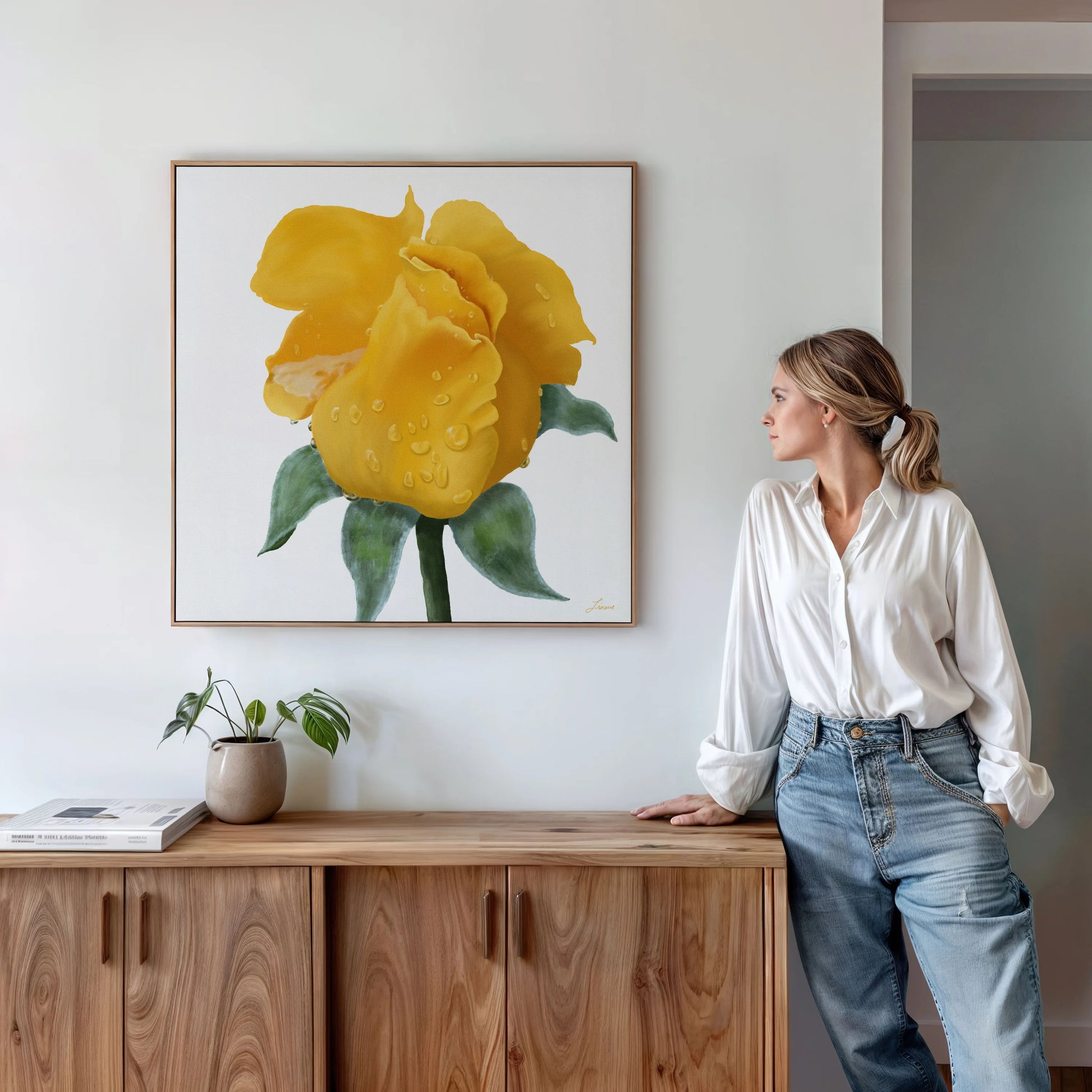 A woman in a white blouse and jeans leans against a wooden sideboard, gazing at a yellow rose canvas art print with water droplets on a white wall—an elegant touch to the contemporary interiors. A small plant and books sit beneath the artwork.