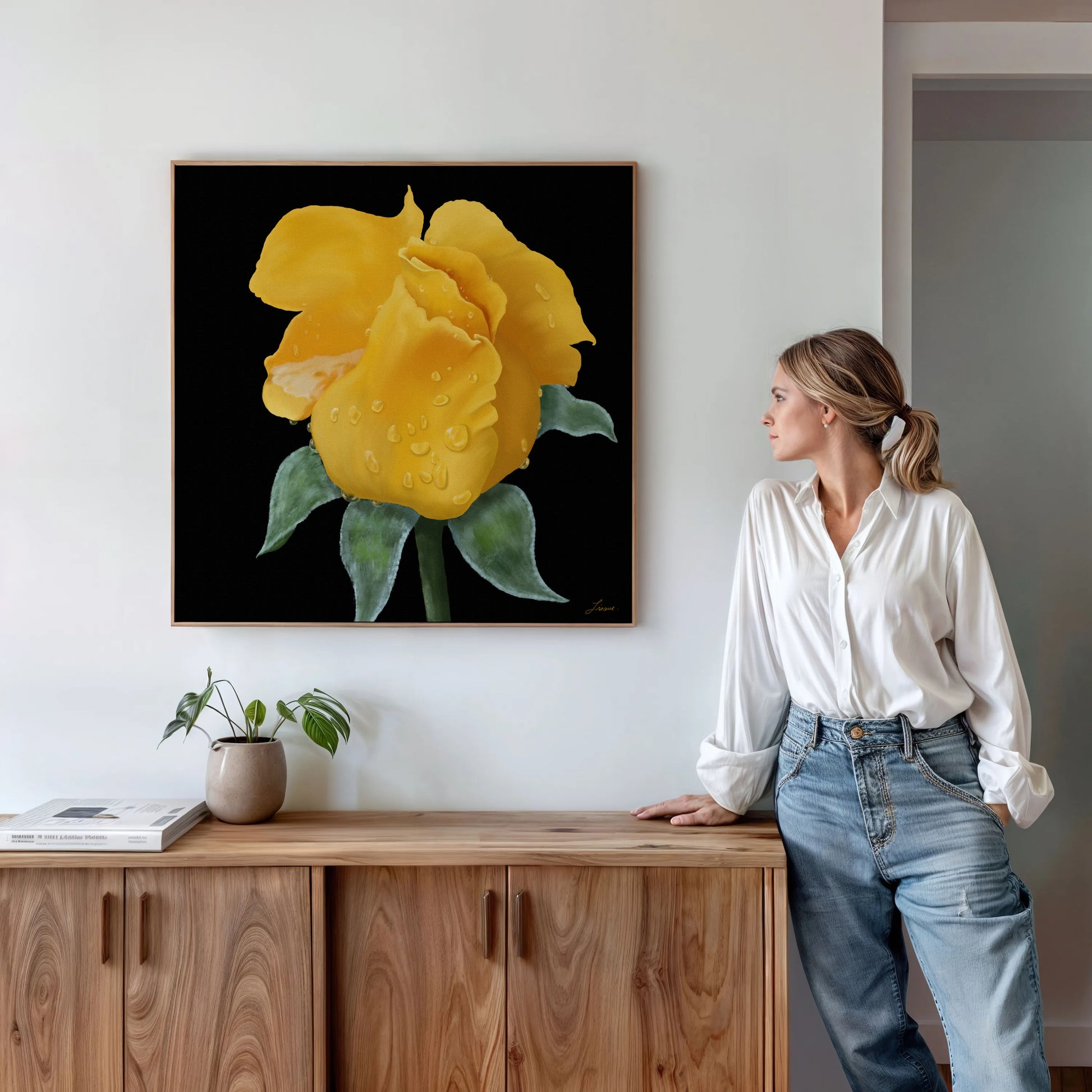 A woman in a white blouse and jeans stands by a wooden cabinet, admiring a canvas art print of a yellow rose with water droplets against a black background—an elegant touch for modern interiors. A potted plant and books complete the scene.