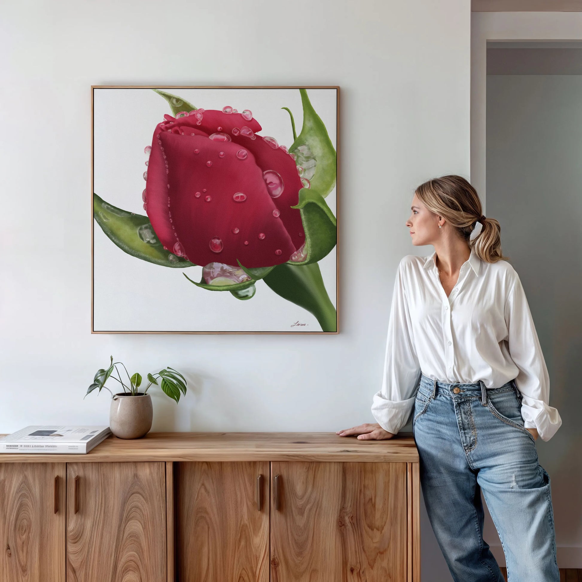 A woman in a white blouse and blue jeans stands by a wooden sideboard, admiring Refreshing Love—a large gallery-wrapped canvas of a red rose with water droplets—hanging on a bright, modern wall. A small potted plant rests nearby.