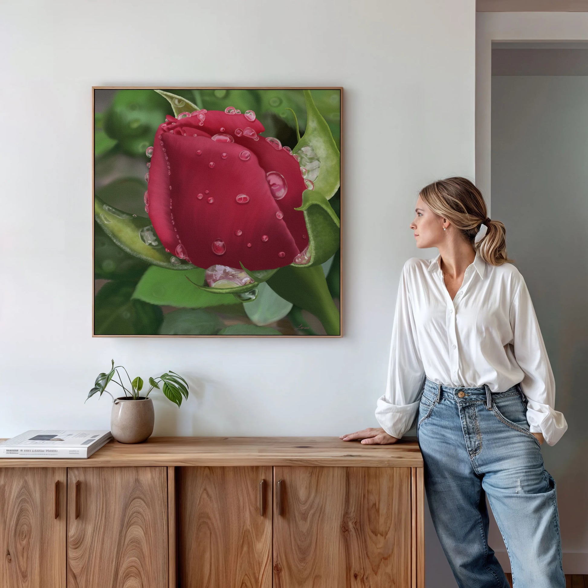 A woman in a white blouse and jeans stands by a wooden sideboard, admiring a romantic wall art red rose canvas with water droplets, displayed on a white wall. A potted plant and books rest nearby, enhancing the inviting scene.