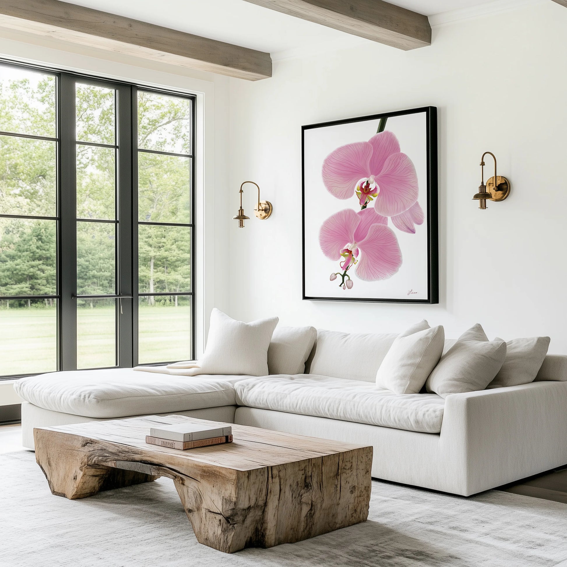A bright living room features a large L-shaped white sofa, wood coffee table, and a book on top. Above the sofa hangs a gallery-wrapped canvas of Pink orchid flowers, flanked by brass sconces. Tall windows overlook a grassy, tree-filled yard.