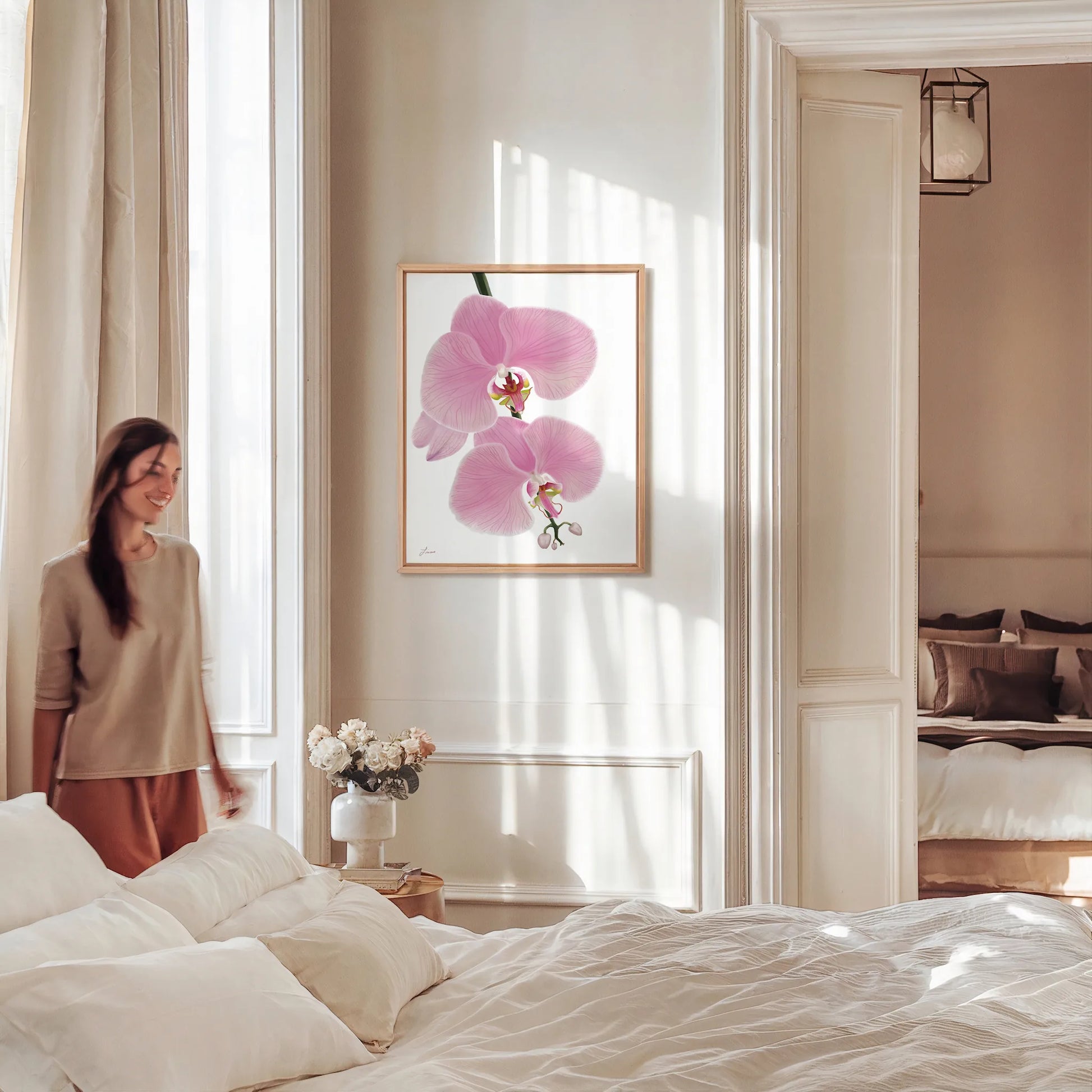 A woman stands smiling near a bright bedroom with neutral decor. Sunlight filters through tall windows. A neatly made bed sits in the foreground, and above it hangs a gallery-wrapped canvas of pink orchid flowers in a floating wood frame. Another bedroom is visible beyond.