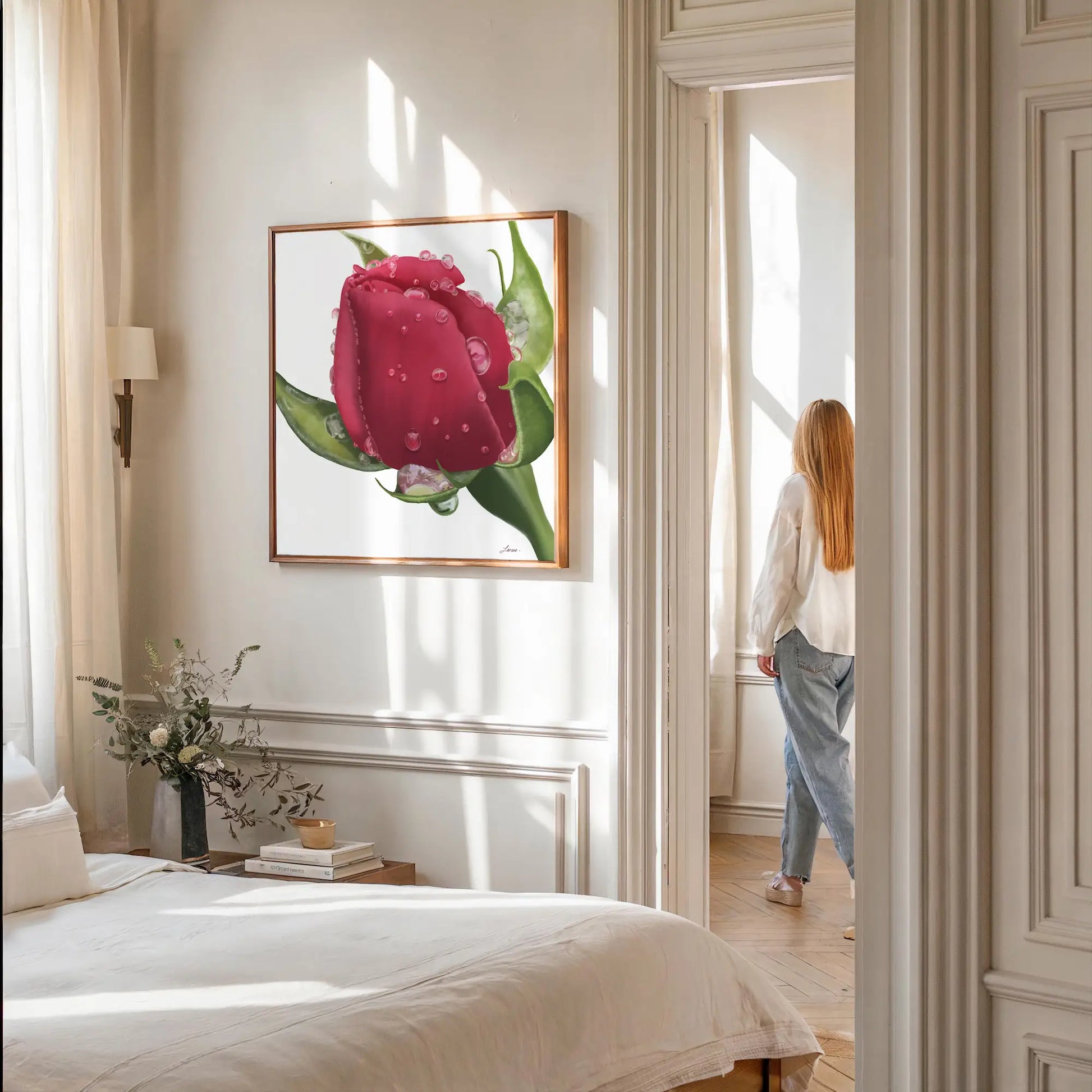 A cozy bedroom with a bed in the foreground and a bedside table holding a vase of flowers and books. A large red rose canvas in a floating wood frame hangs on the wall, while in the background, a woman gazes out from a sunlit room.