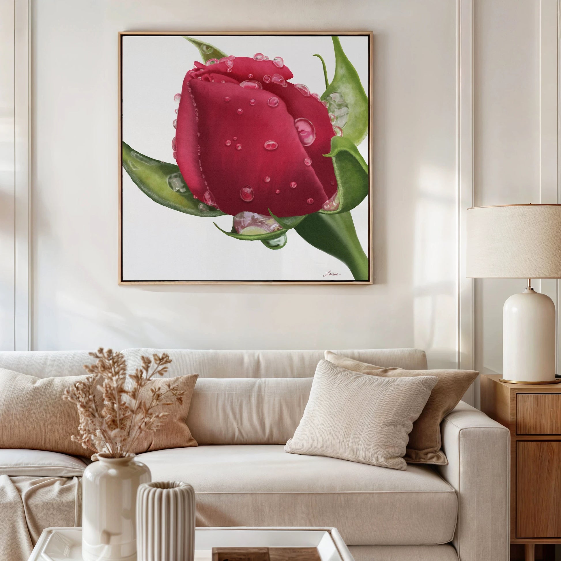 A living room with a white sofa, beige cushions, and a light wooden side table with a white lamp. A gallery-wrapped canvas depicting a red rosebud with water droplets and green leaves adds elegance above the sofa.