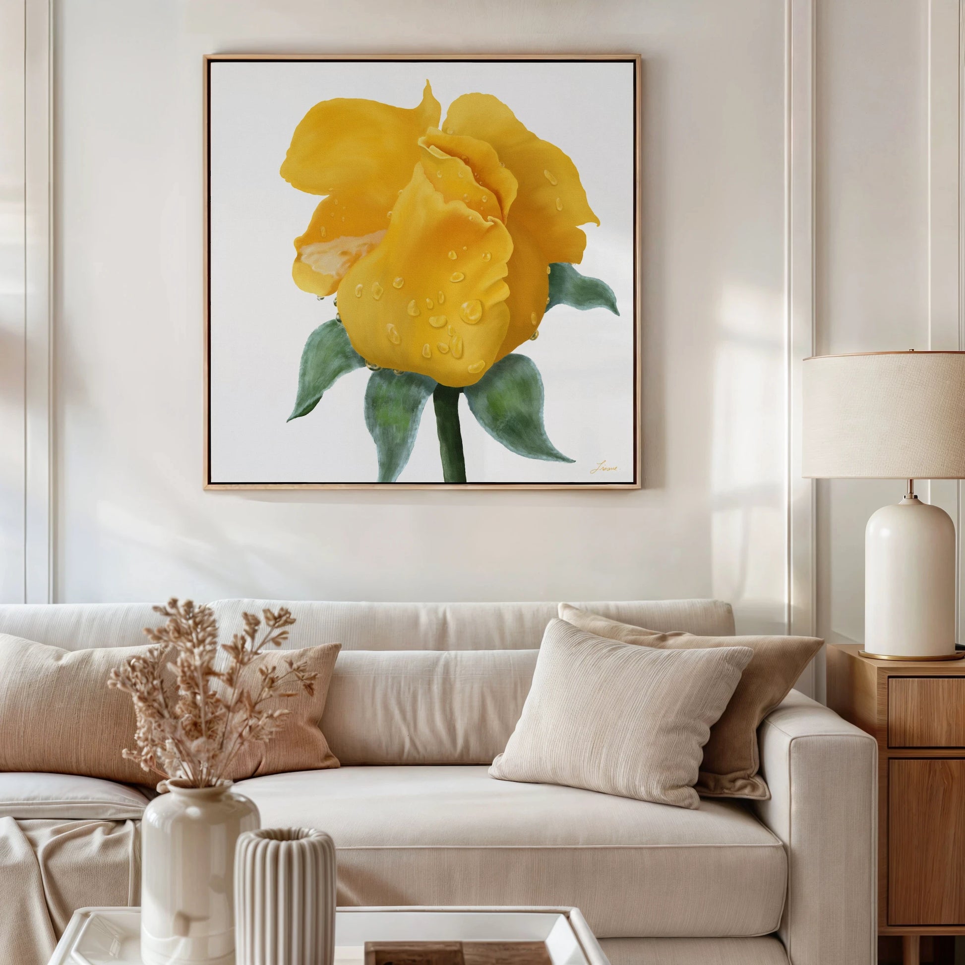 A modern living room with neutral tones features a beige sofa, cream pillows, dried flowers in a white vase, a table lamp, and a yellow rose gallery-wrapped canvas with water droplets and green leaves above the sofa.