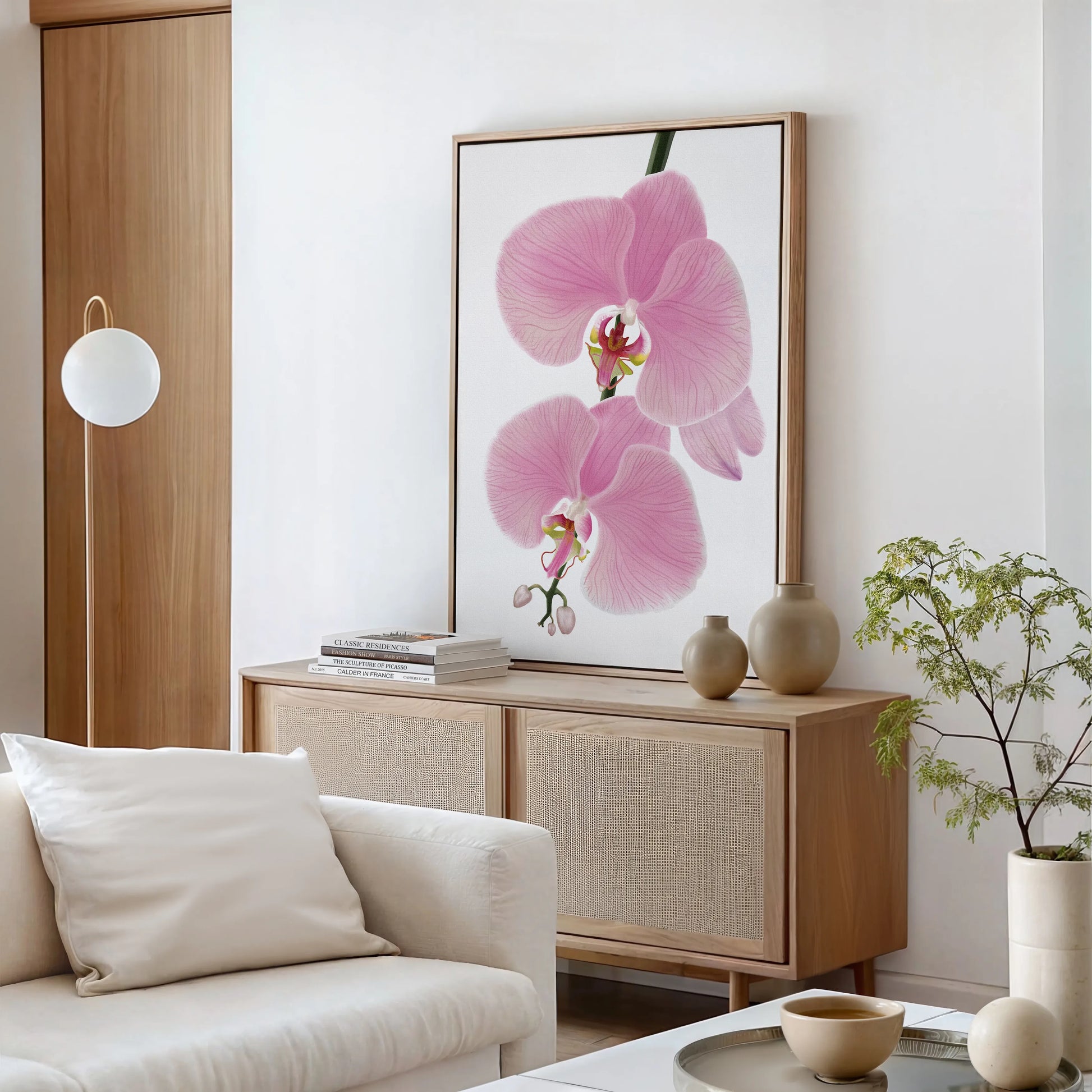 A modern living room with a light wood sideboard, beige vases, stacked books, a gallery-wrapped canvas of pink orchid flowers in fade-resistant color, a leafy plant, a round lamp, and a white sofa; decor is minimalist and neutral-toned.