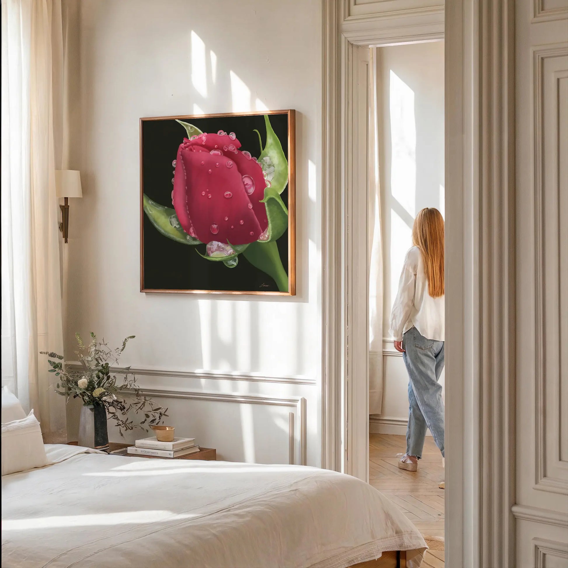A softly lit bedroom with white walls features a bed with neutral bedding, a wooden nightstand holding a vase of white flowers, and a large hyperrealistic red rose on gallery-wrapped canvas. A person with long hair stands by a bright window in the adjoining room.